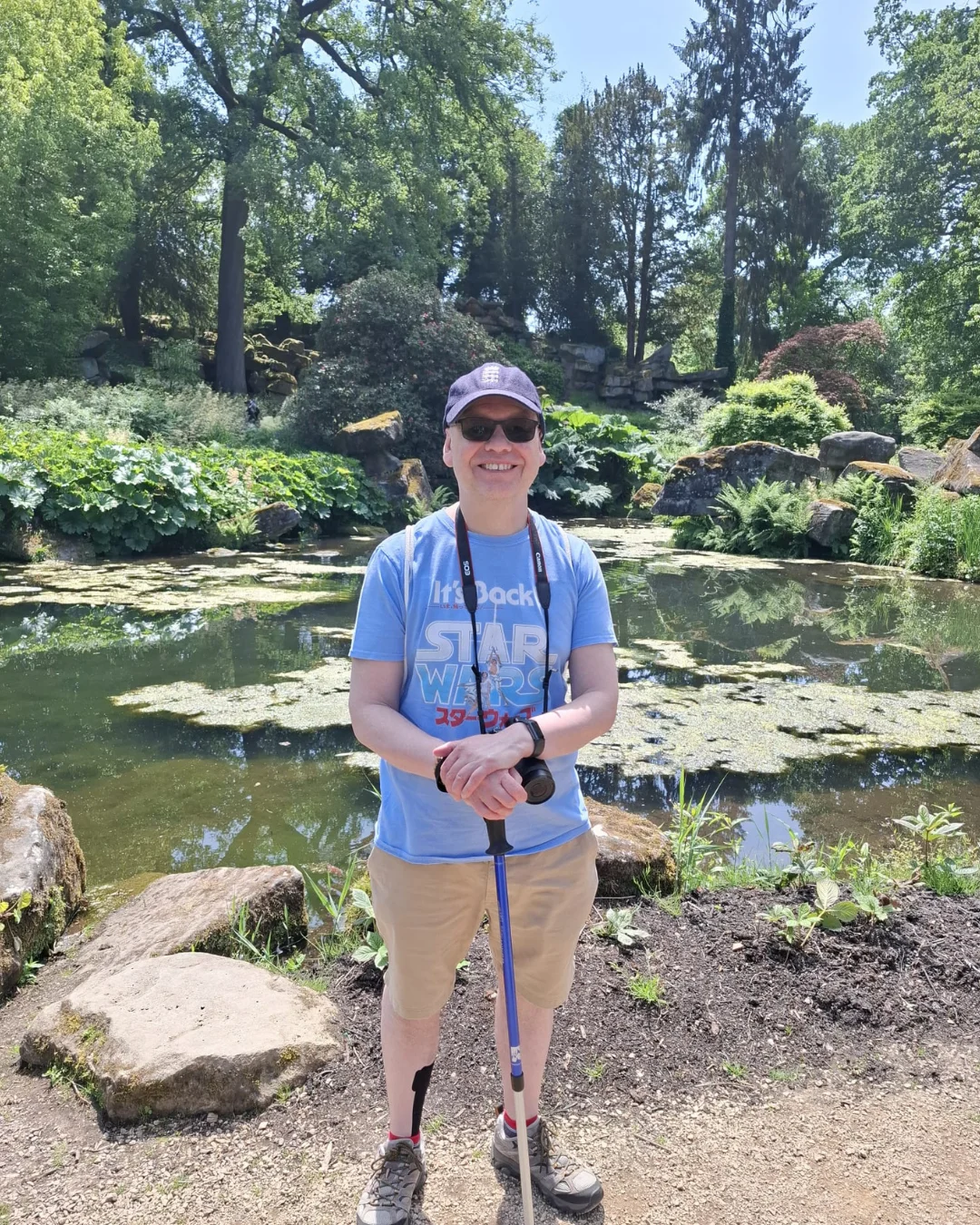 A man stands in front of a garden and pond smiling and holding a walking stick