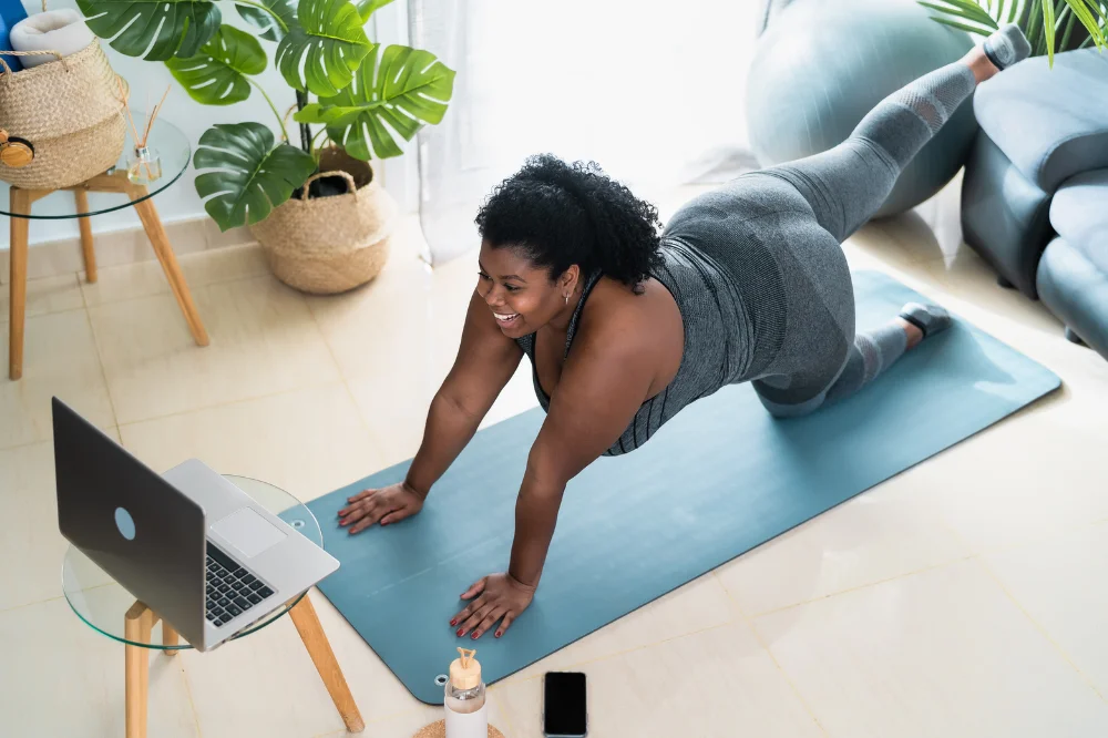 Woman on a yoga mat in a living room, on all fours, lifting one leg into the air during a bird dog exercise
