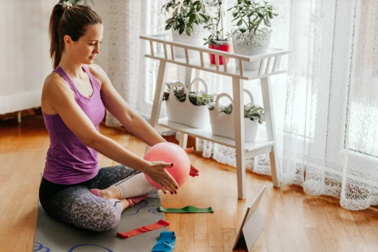 Woman does exercise program and squeezes a pilates ball and sits on a mat looking at a tablet screen
