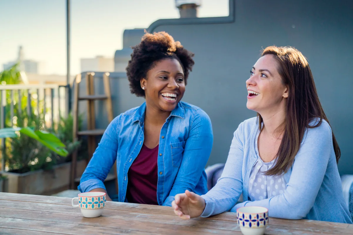 Two women laughing together over coffee outdoors, representing the connection members find in The Zebra Club