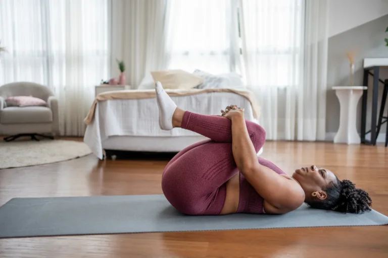 Woman doing a gentle knees-to-chest stretch on a yoga mat, an example of low-exertion movement suited to hypermobility and ME/CFS pacing.