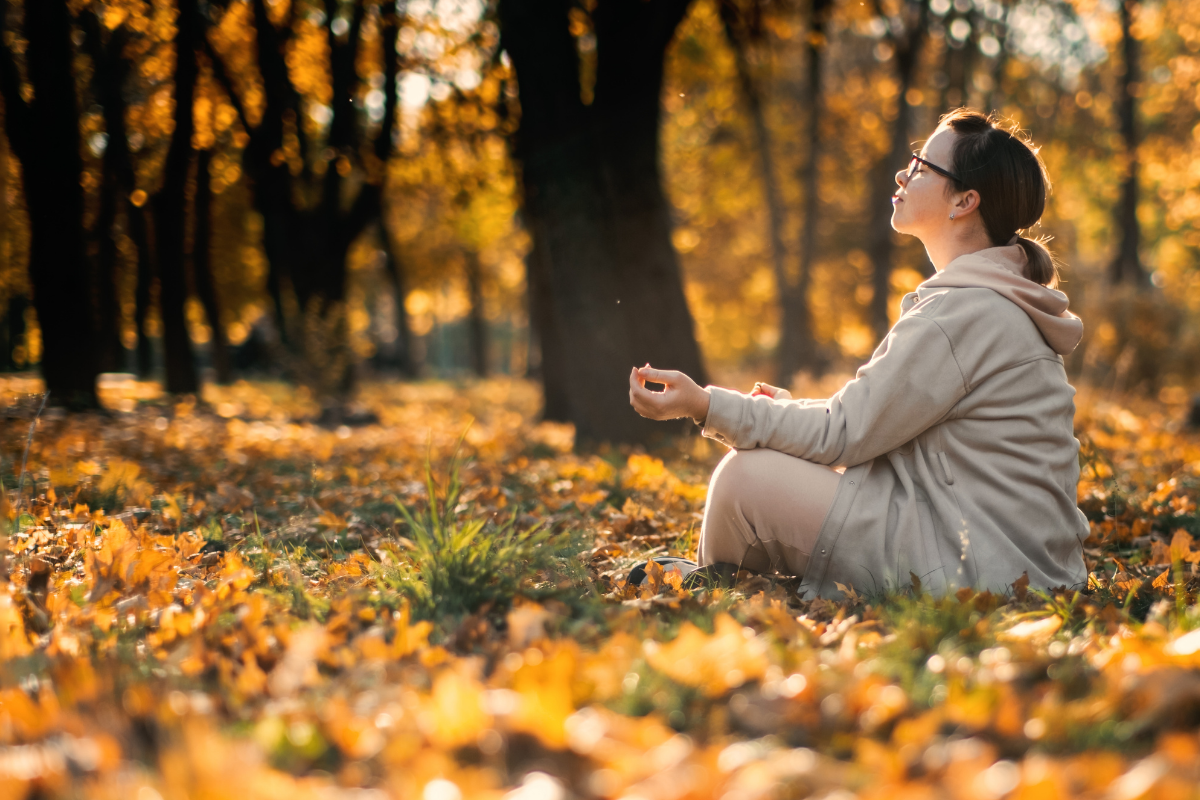 Woman sitting cross-legged among golden leaves, eyes closed and face turned toward the sun