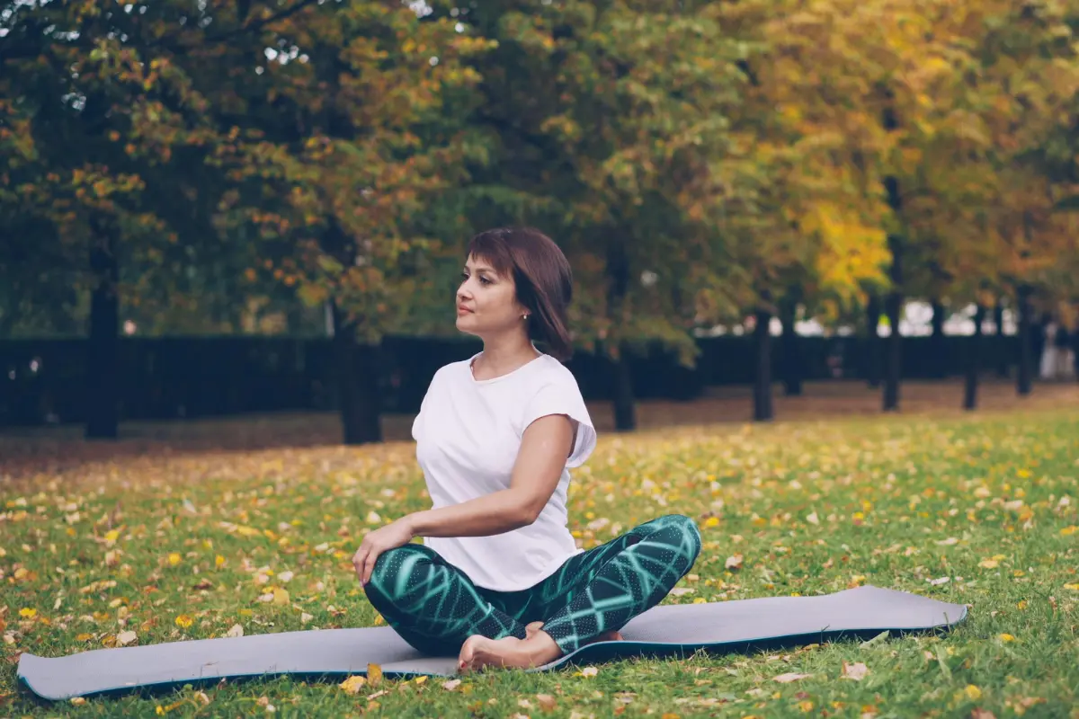 Woman seated on a yoga mat in a park, cross-legged in a gentle twist among autumn leaves