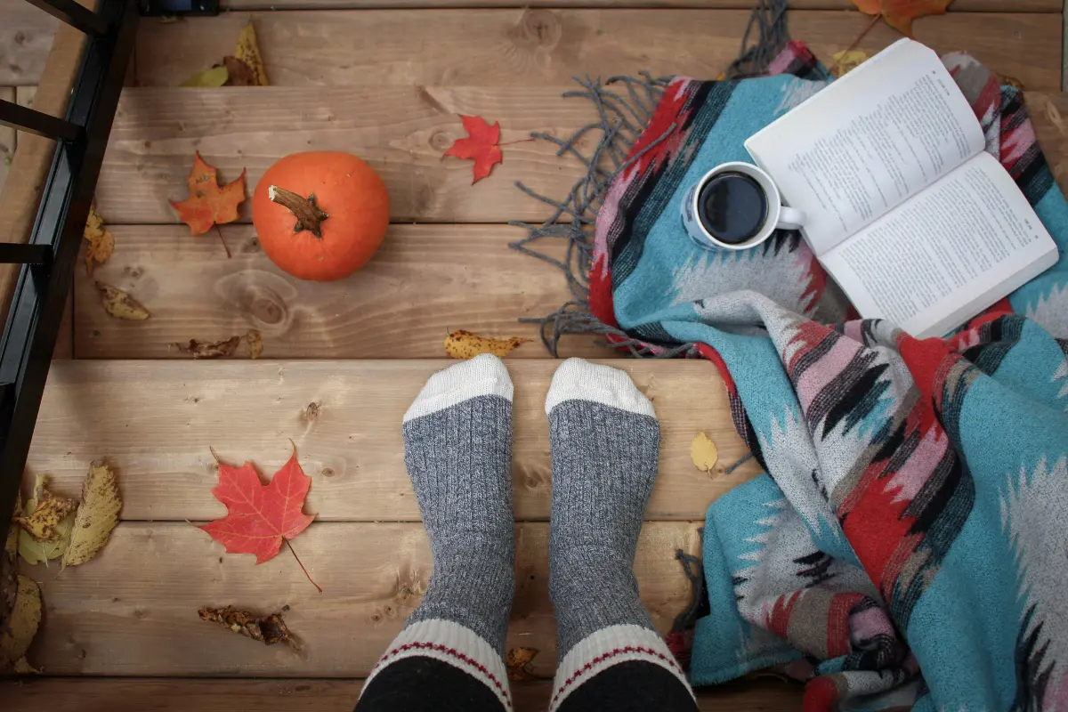 Wool socks on a wooden porch beside a small pumpkin, fallen leaves, a book, coffee, and a blanket