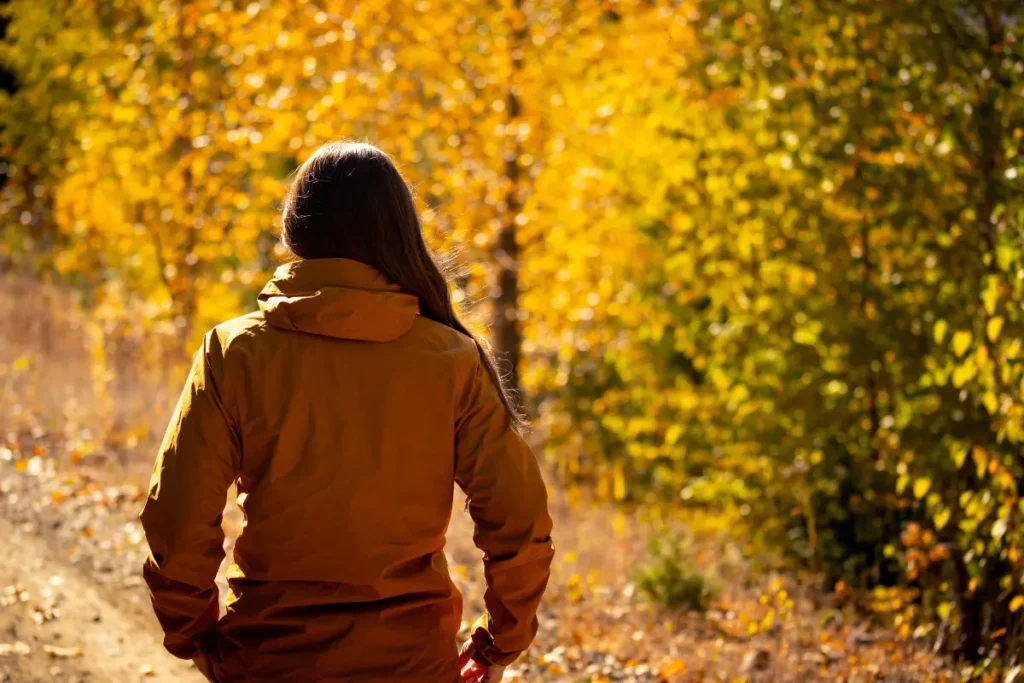 A woman walks alone on an autumn path through golden trees, illustrating the October Slide in chronic illness